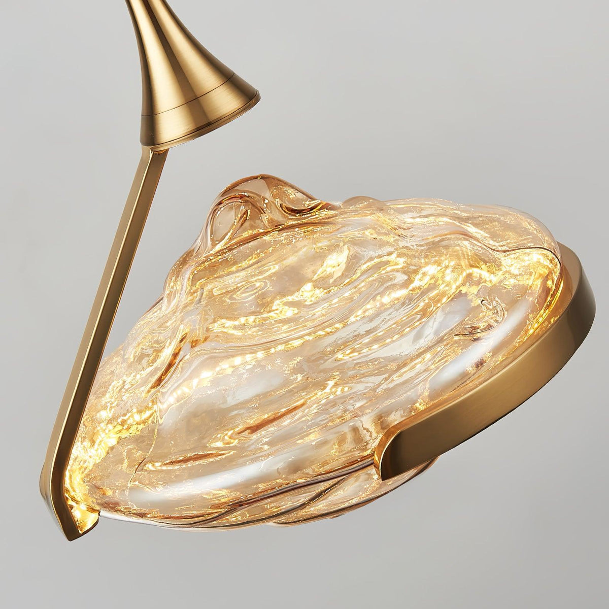 Close view of the rippled amber glass shade, lit within, and its gold ring and curved arm on this Clérachic pendant.