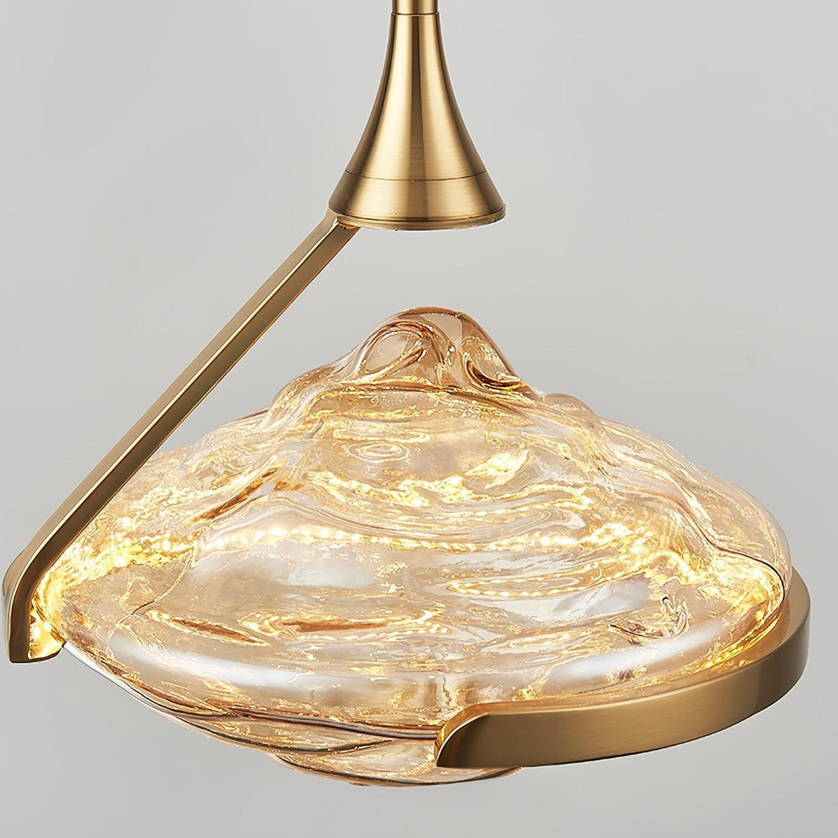 A faceted amber glass shade glows beneath a brushed gold flared rod joint against a pale grey backdrop, a Clérachic pendant.