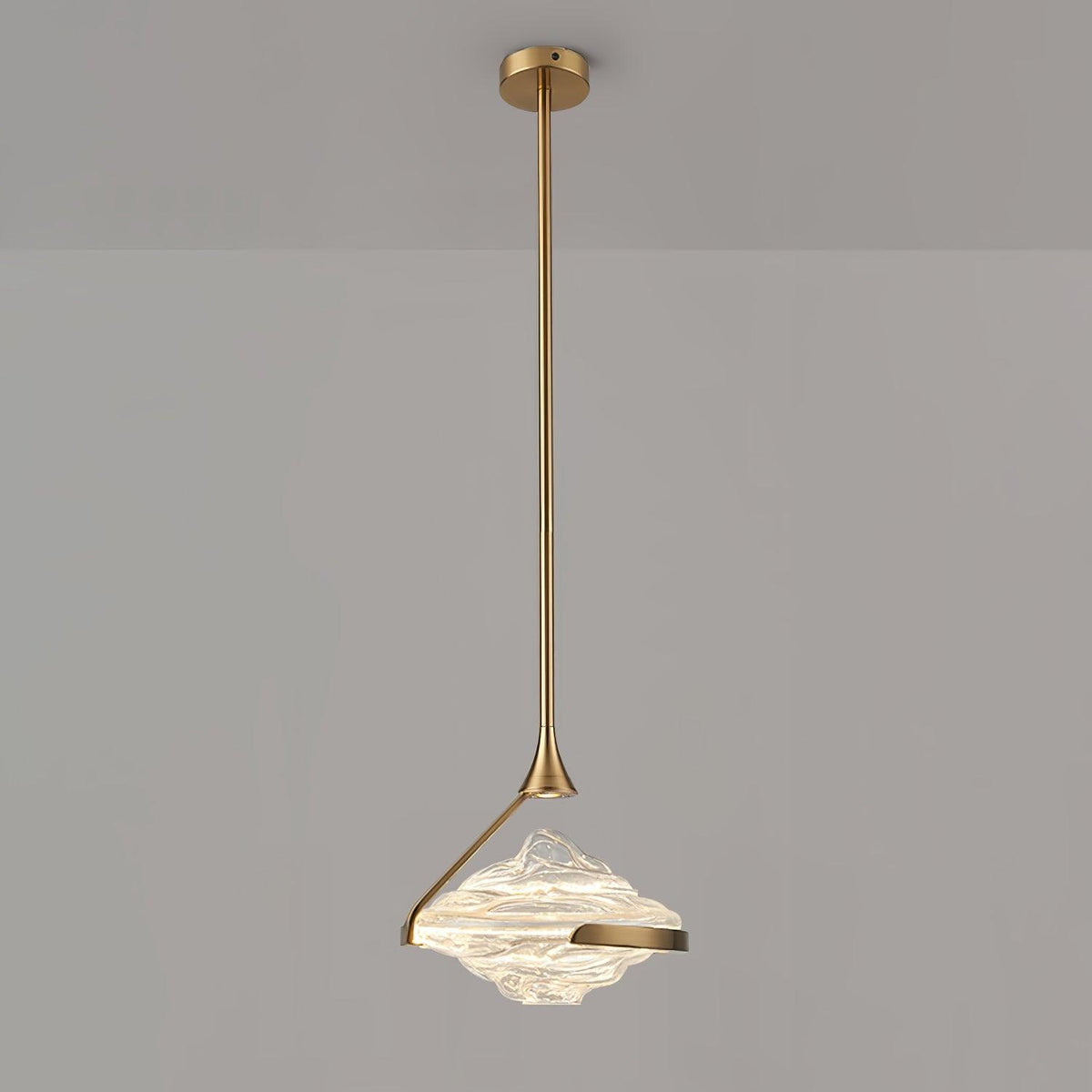A faceted clear glass pendant with a gold arm and rod hangs against a grey wall, lit white, from Clérachic.
