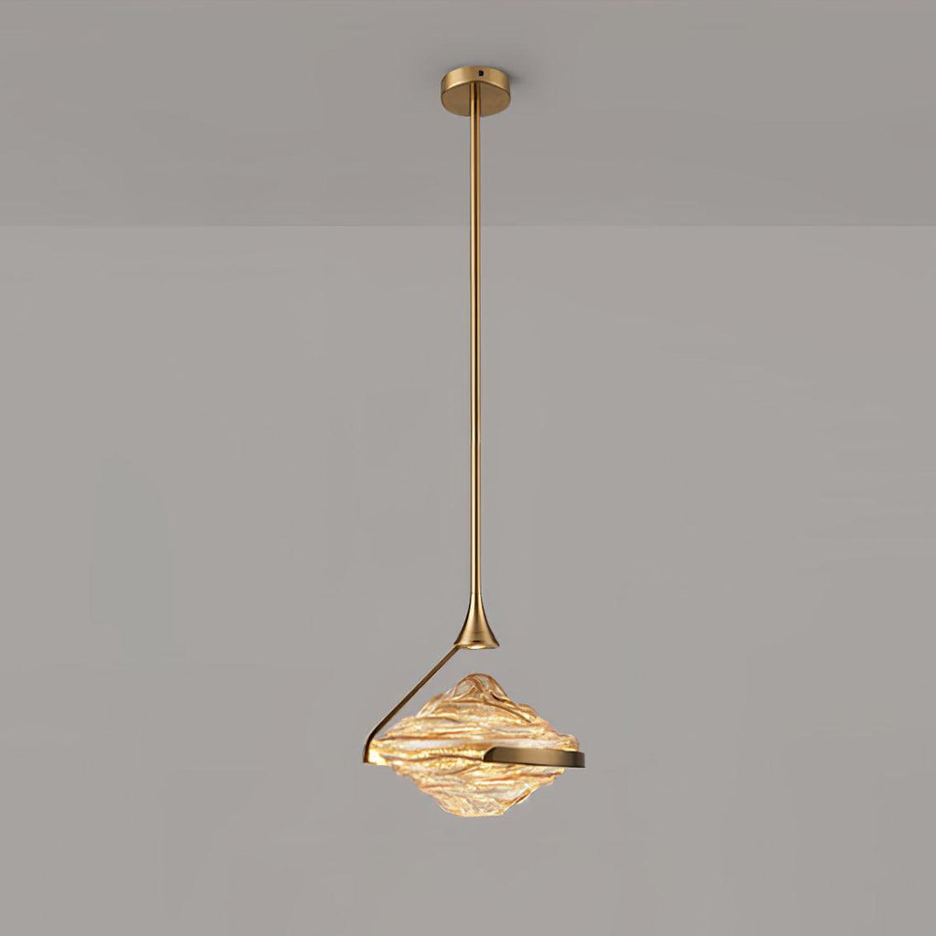 A faceted amber glass pendant with a gold arm and ceiling plate hangs against a grey wall, glowing warm, from Clérachic.