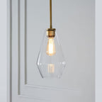 Clérachic faceted glass pendant light in clear glass, the slim teardrop shade lit beside a white panelled wall.