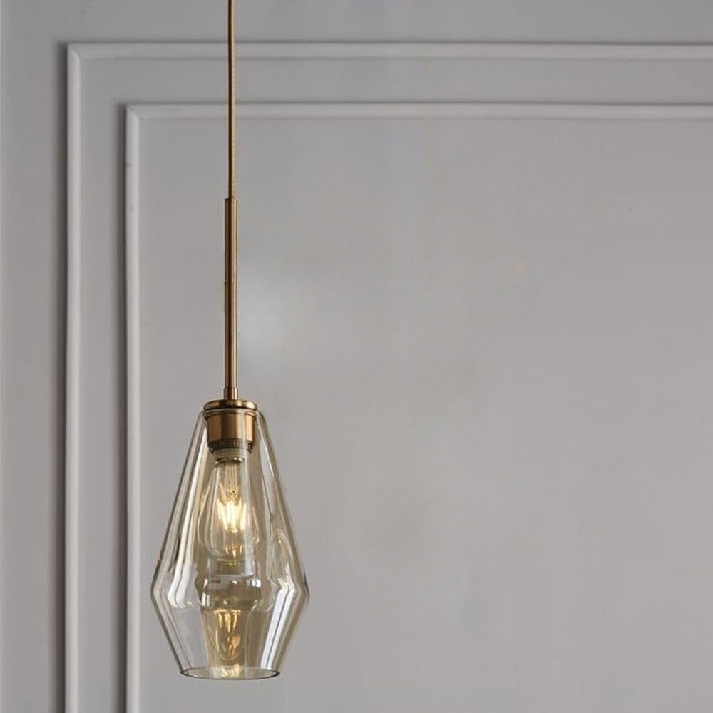 The amber elongated teardrop of the Clérachic faceted glass pendant light on its brass-tone rod against a grey wall.
