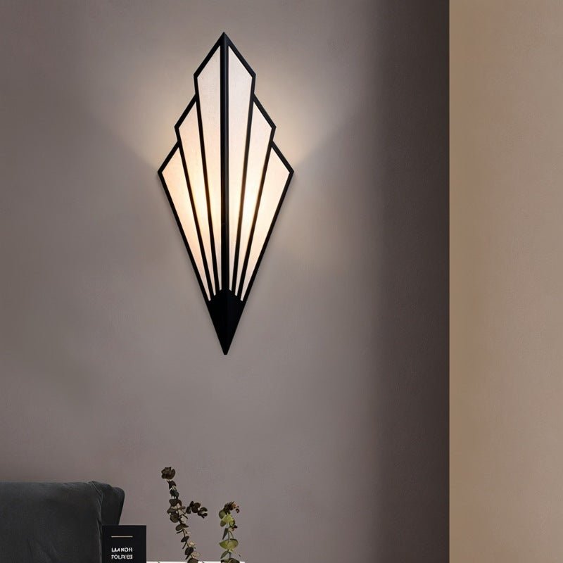 Clérachic black Art Deco wall lamp with fan-shaped linen panels lit warm on a taupe wall above a sofa arm and a plant stem.