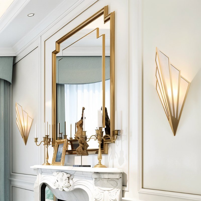Clérachic gold fan-shaped wall lamps lit warm on a panelled white wall beside a gold-framed mirror above a marble fireplace.