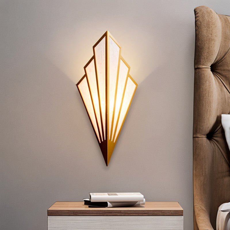 Clérachic gold Art Deco wall lamp glowing warm beside a tufted headboard, above a bedside table with a stack of books.