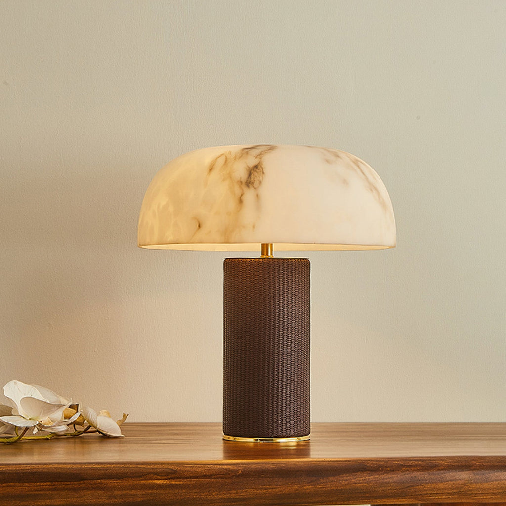 A Clérachic table lamp with a glowing domed marbled shade and a dark woven base on a wooden table, with white orchids beside it.