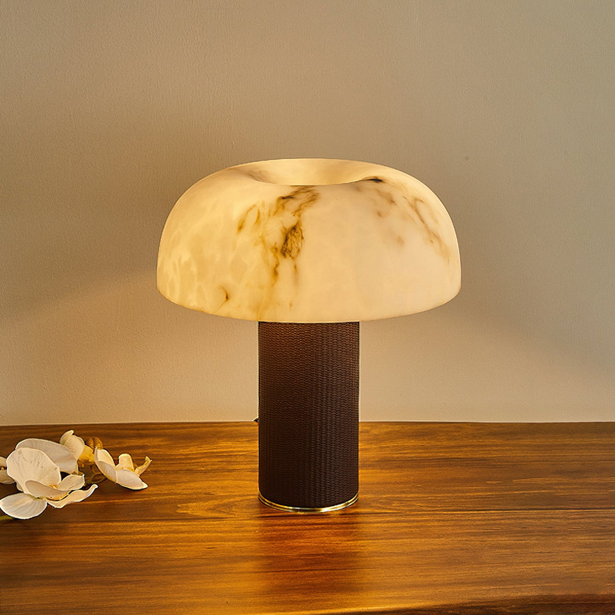 Slightly raised view of a Clérachic table lamp lit, with a softly glowing mushroom-shaped shade and a dark woven base.