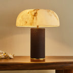 Clérachic table lamp lit warm, with a wide domed shade, brass-toned trim and a dark base, on a wooden table.