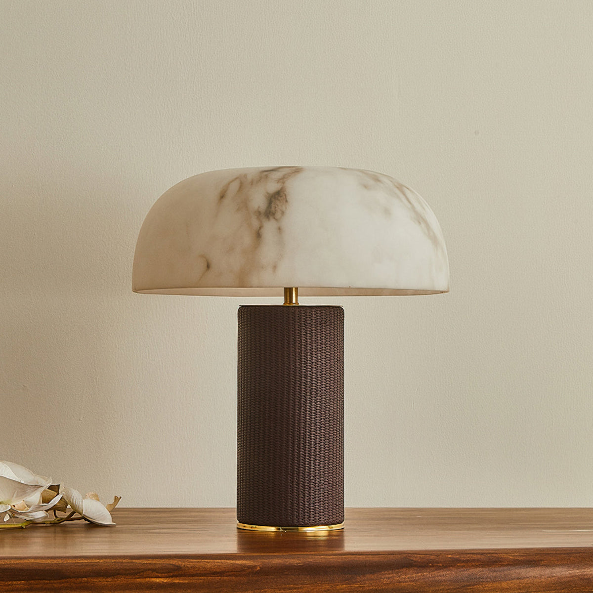 Unlit Clérachic table lamp with a marbled white shade and a dark woven base, on a wooden table against a cream wall.
