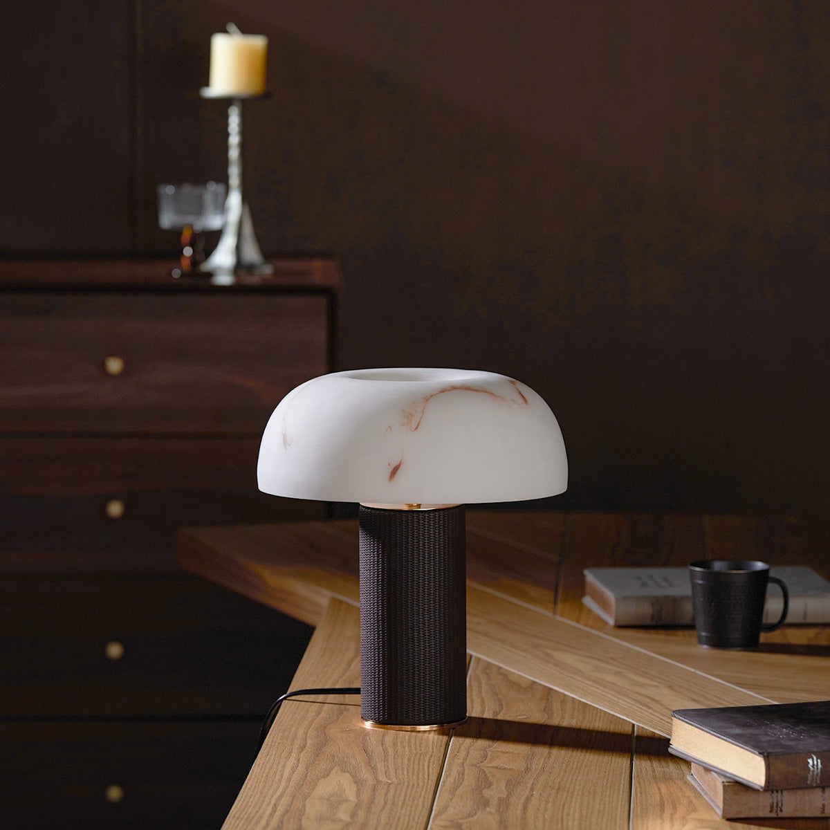 An unlit Clérachic table lamp stands on a pale wood desk in a dark room, with books, a mug and a candlestick behind.