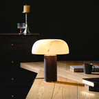 Lit Clérachic table lamp glowing in a dim room on a light wood desk, with books and a dark mug beside it.