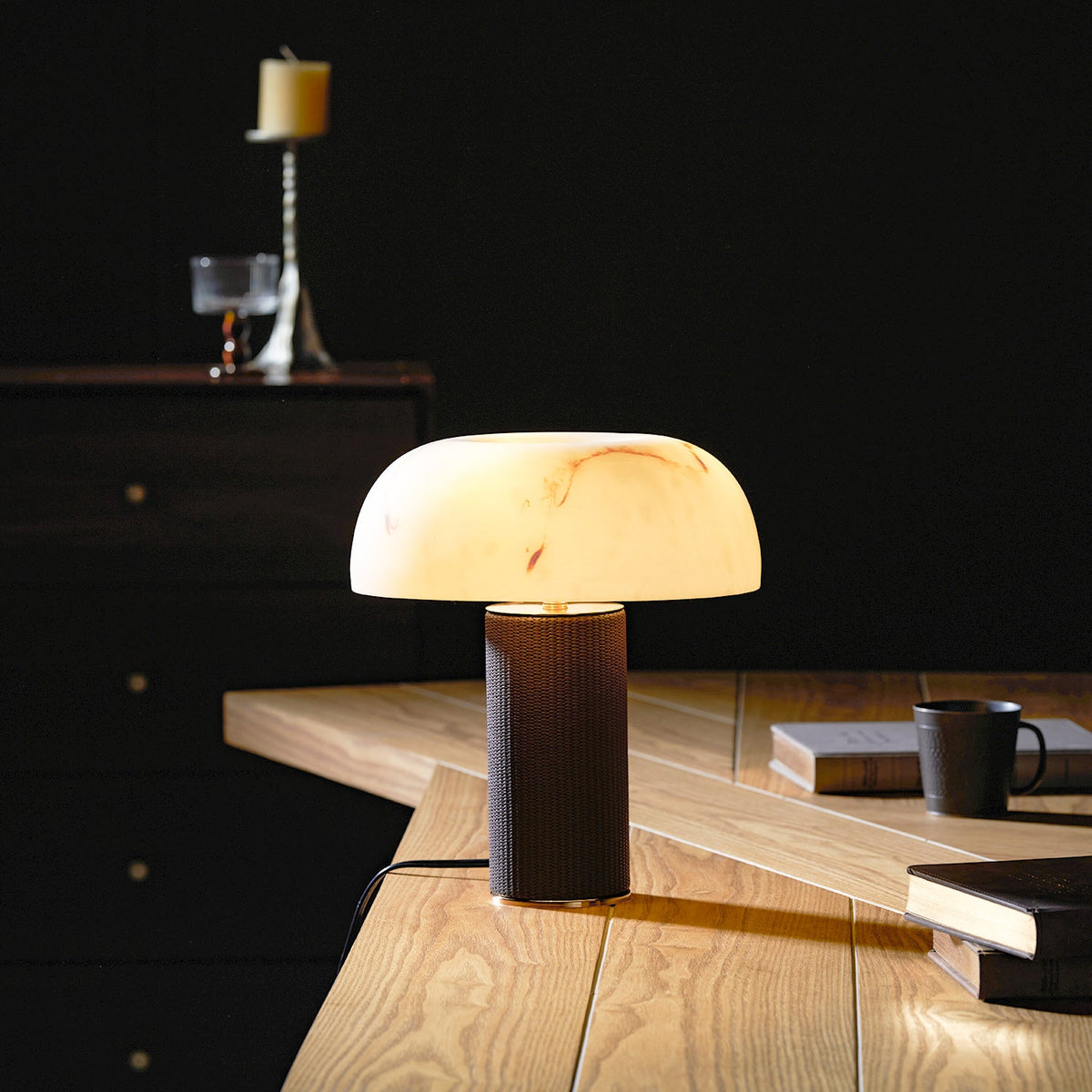 Lit Clérachic table lamp glowing in a dim room on a light wood desk, with books and a dark mug beside it.