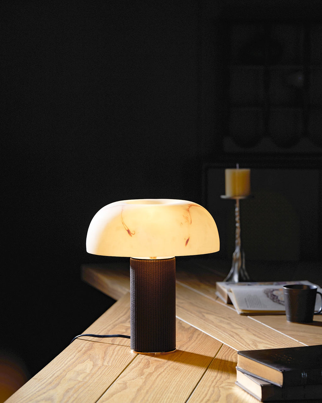 A Clérachic table lamp lights a corner of a pale wood desk in a dark room, with a magazine and a candle in the background.