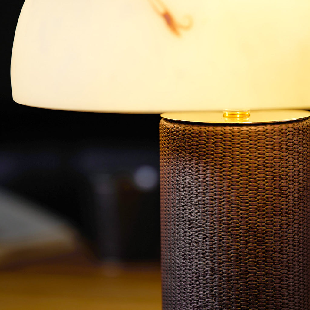 Close view of a lit Clérachic table lamp, with its glowing shade above the woven base, on a dark blurred background.
