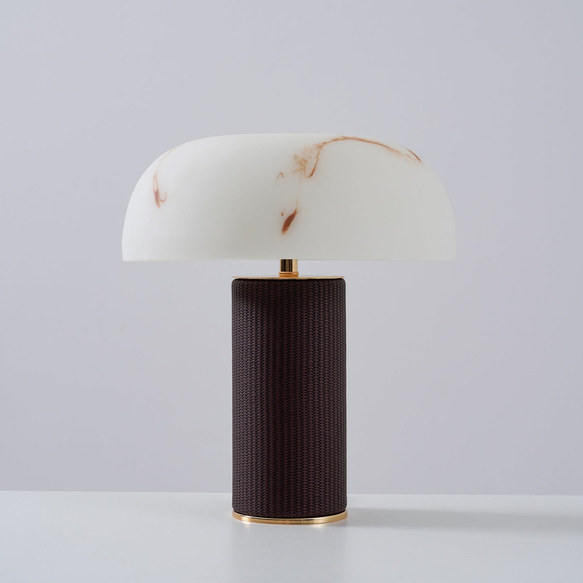 Unlit Clérachic table lamp with a white veined shade and a dark woven base with a gold ring, on a light backdrop.