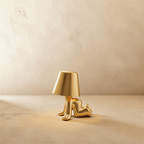 Clérachic gold figurine table lamp reclining on its side, warm glow beneath a tapered shade against a plaster wall.