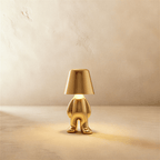 Clérachic gold figurine table lamp standing upright, warm glow beneath its tapered shade against a plaster wall.