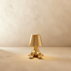 Clérachic gold figurine table lamp seated with hands aglow beneath its tapered shade against a plaster wall.
