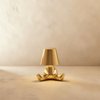 Clérachic gold figurine table lamp seated cross-legged, warm glow beneath its tapered shade against a plaster wall.