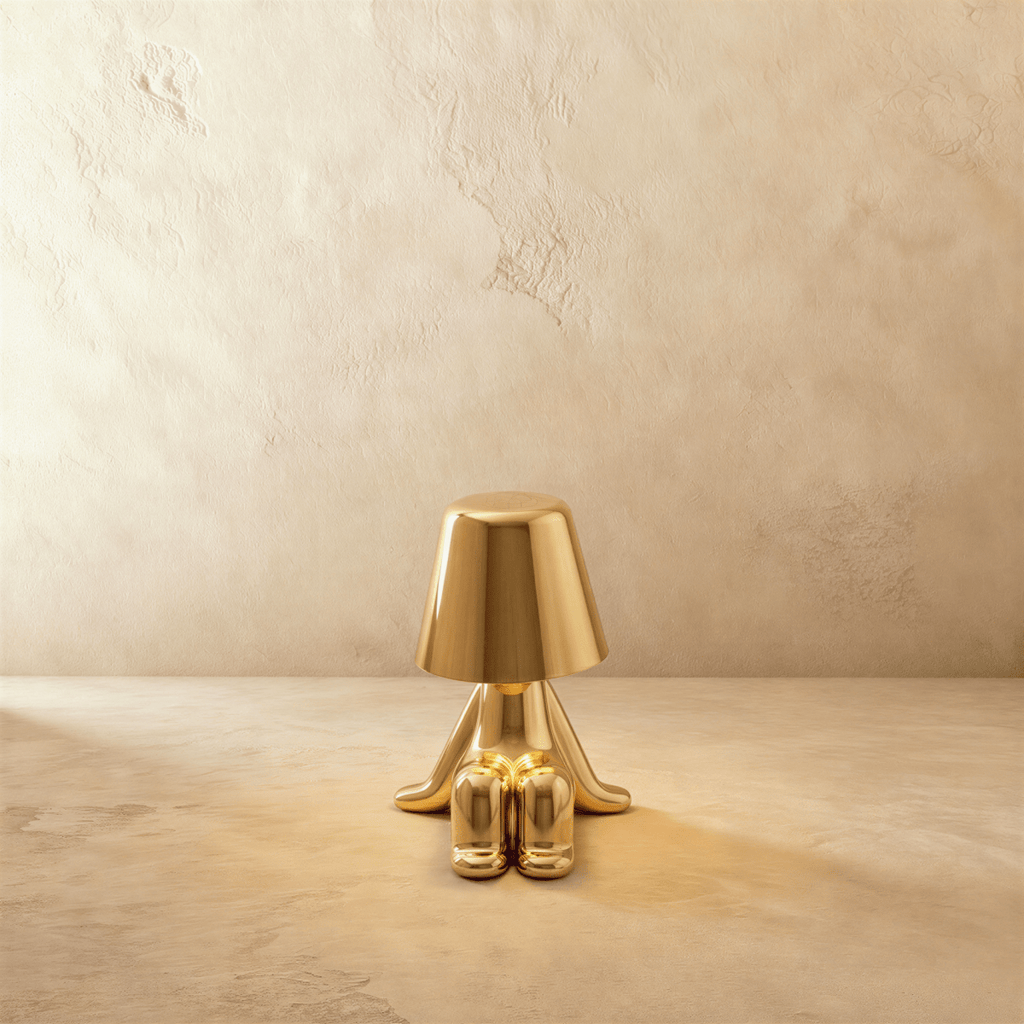 Clérachic gold figurine table lamp seated with feet crossed, warm glow beneath its tapered shade against a plaster wall.