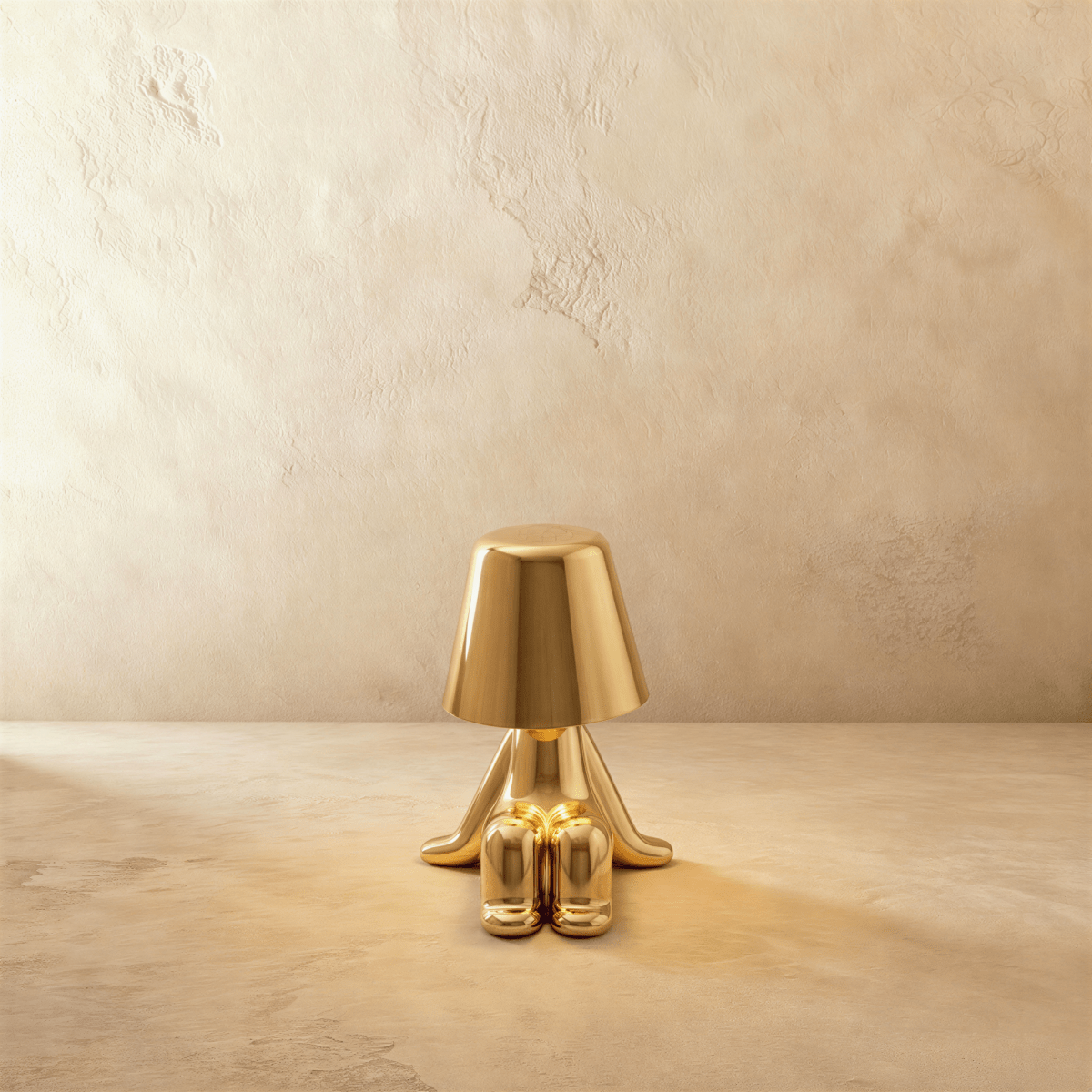 Clérachic gold figurine table lamp seated with feet crossed, warm glow beneath its tapered shade against a plaster wall.