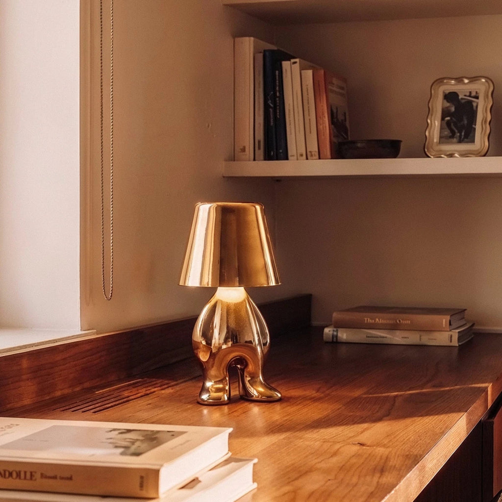Clérachic gold figurine table lamp glowing warm white on a wooden shelf beside stacked books and a framed photo.