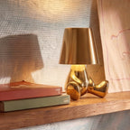Clérachic gold figurine table lamp glowing warm white on a wooden shelf beside books and a child's pencil drawing.