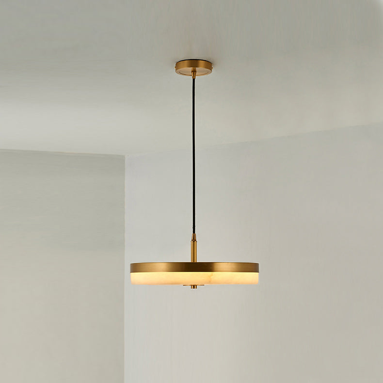 The brass-rim Clérachic Flat Disc Pendant Light lit in a bare white corner, its alabaster edge glowing warm beneath the metal band.