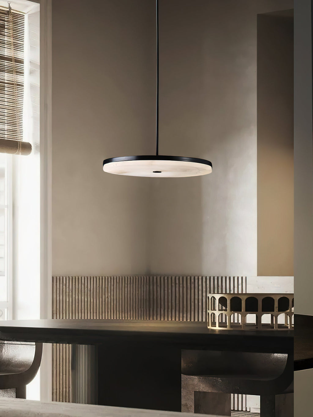 Clérachic Flat Disc Pendant Light in black, lit above a dark dining table in a plaster-walled room with a bamboo blind at the window.