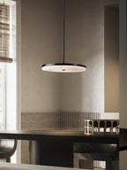 Clérachic Flat Disc Pendant Light in black, lit above a dark dining table in a plaster-walled room with a bamboo blind at the window.