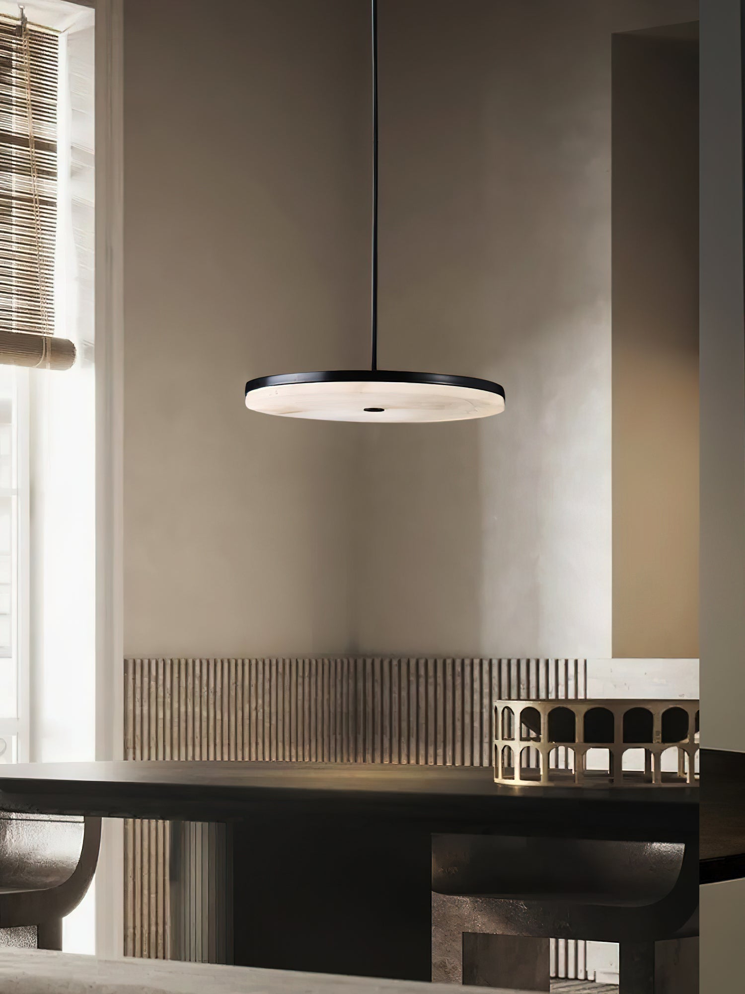 Clérachic Flat Disc Pendant Light in black, lit above a dark dining table in a plaster-walled room with a bamboo blind at the window.