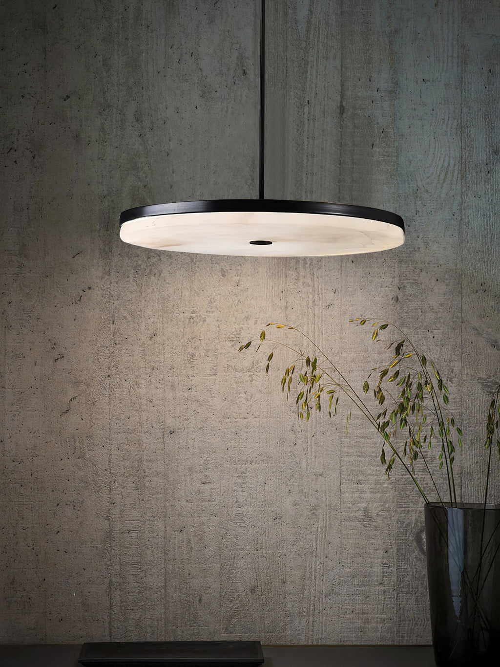 The Clérachic Flat Disc Pendant Light against a raw concrete wall, its downward halo lighting dried grasses in a glass vase below.