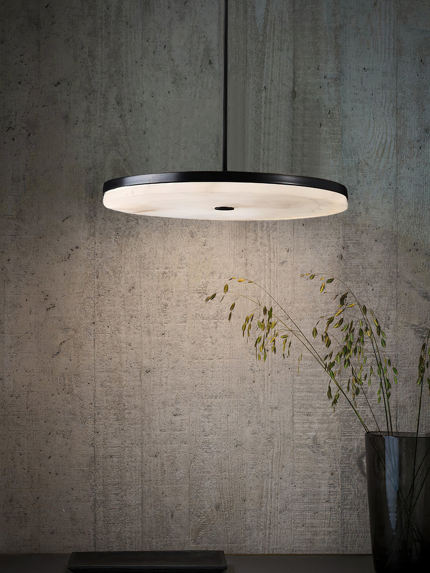 The Clérachic Flat Disc Pendant Light against a raw concrete wall, its downward halo lighting dried grasses in a glass vase below.