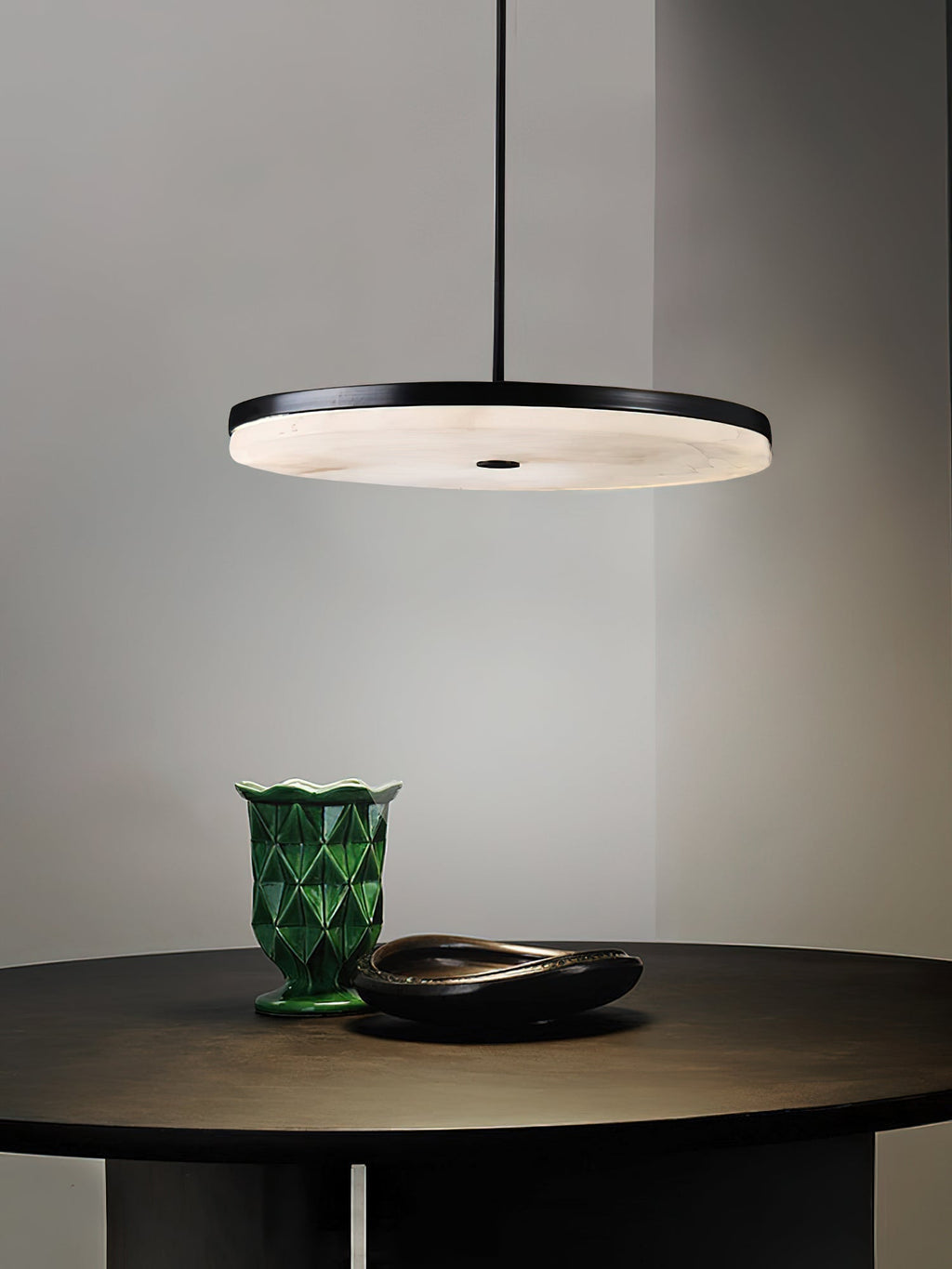 Clérachic Flat Disc Pendant Light lighting a bronze table set with a green ceramic vase, the alabaster underside glowing honey warm.