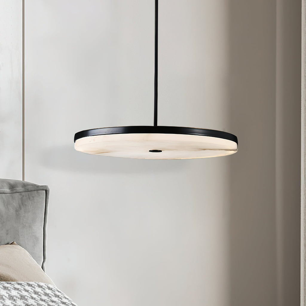 Clérachic Flat Disc Pendant Light lit at bedside height beside a grey velvet headboard, alabaster face bright under a black rim.
