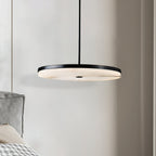 Clérachic Flat Disc Pendant Light lit at bedside height beside a grey velvet headboard, alabaster face bright under a black rim.
