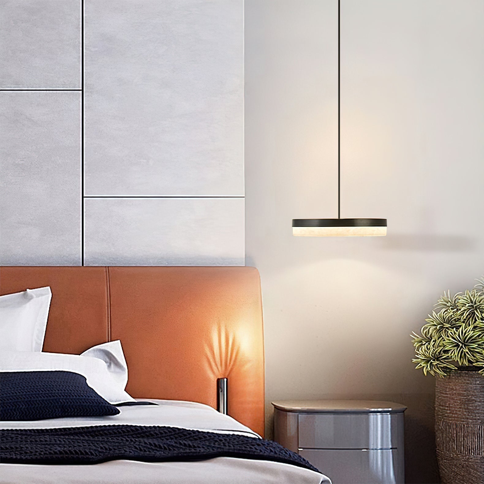 Clérachic Flat Disc Pendant Light on a long drop cord beside a tan leather bed, a warm pool of light on the wall behind it.