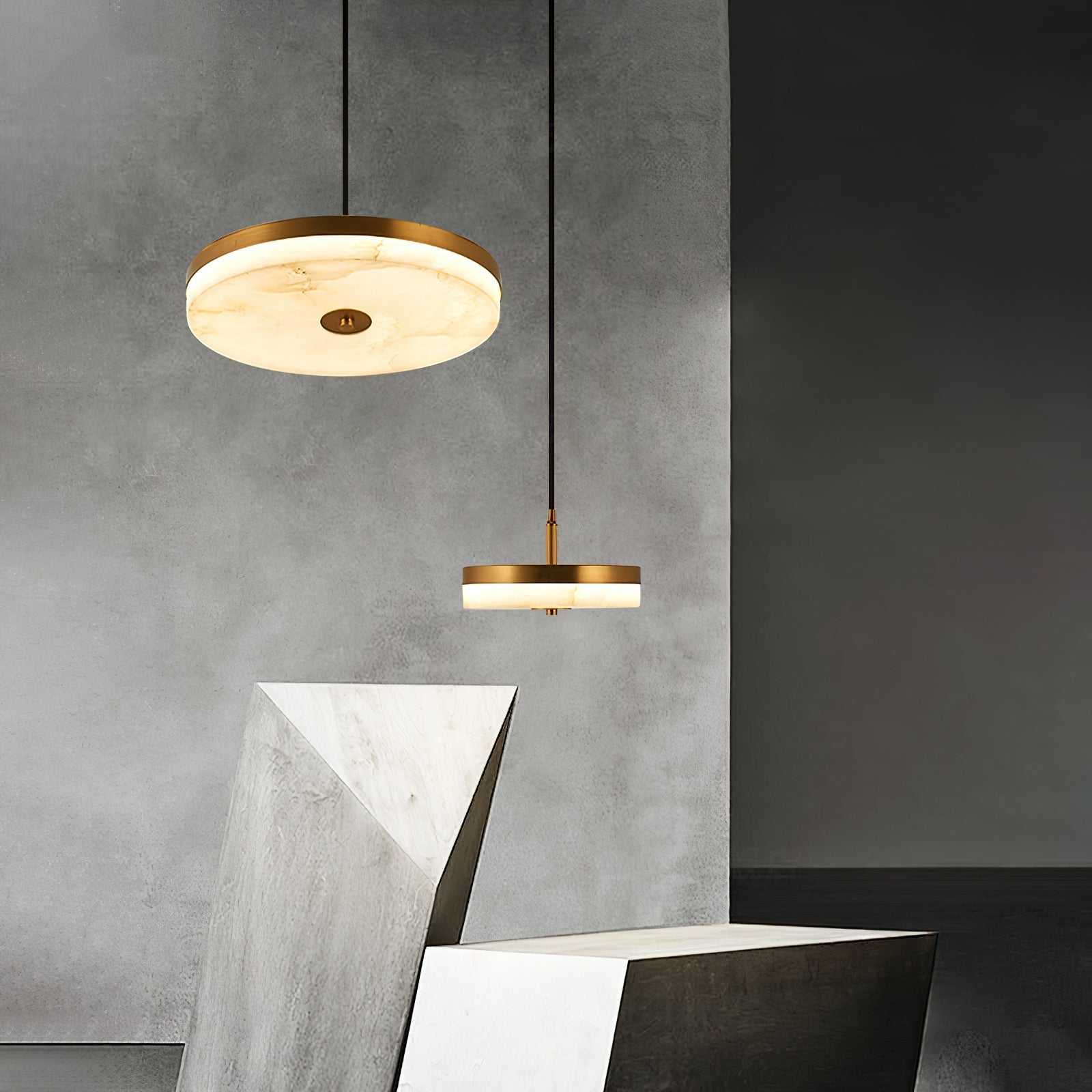 Two brass Clérachic Flat Disc Pendant Light units hung at staggered drops against a concrete wall above pale stone plinths.