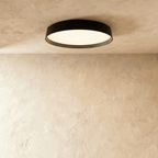 Clérachic flat round ceiling light with a black rim and a diffused white panel glowing softly, mounted on a plaster ceiling.