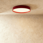 Clérachic flat round ceiling light with a deep red rim and orange inner edge, its diffuser glowing on a plaster ceiling.