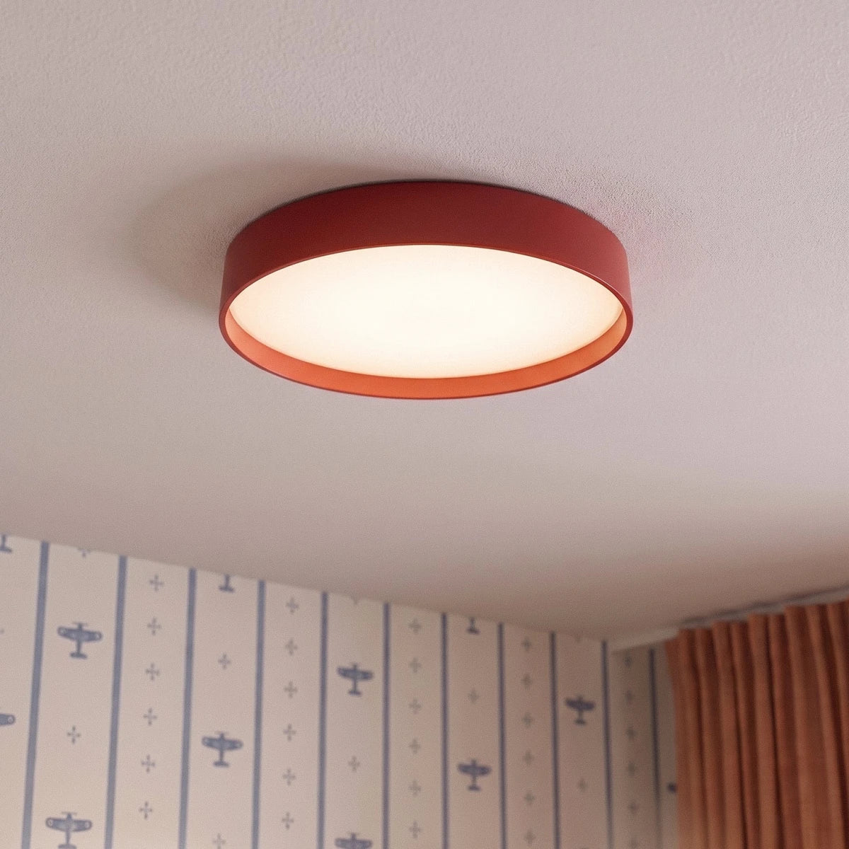 Close view of a Clérachic red round ceiling light with a lit white diffuser, beside aeroplane-print wallpaper and rust curtains.