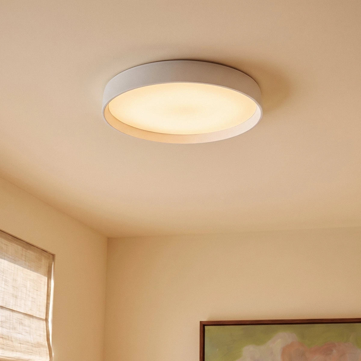 Close view of a Clérachic white round ceiling light, its diffuser glowing warm, beside a linen blind and a framed painting.