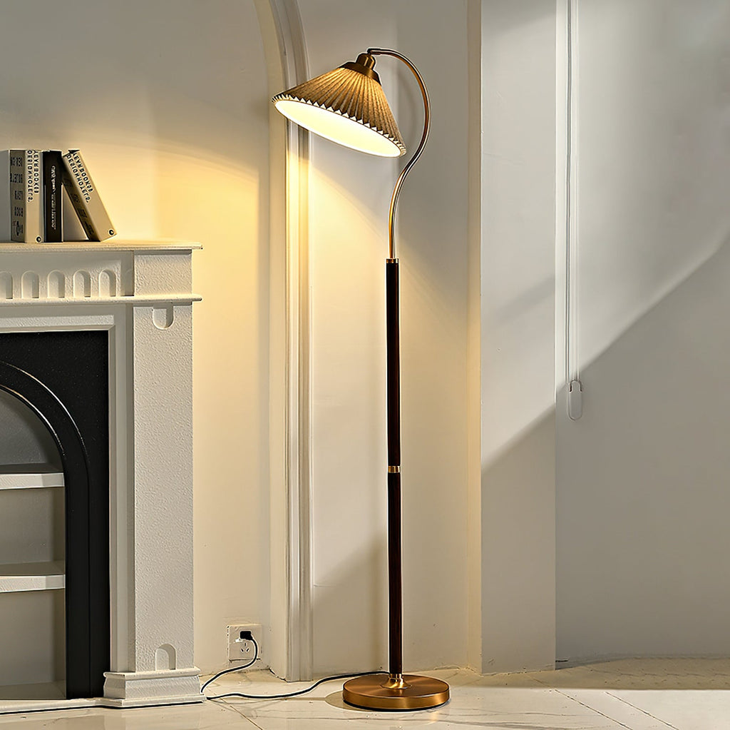 A lit Clérachic floor lamp with a pleated shade curves out beside a white fireplace and shelves of books, light pooling on the wall.