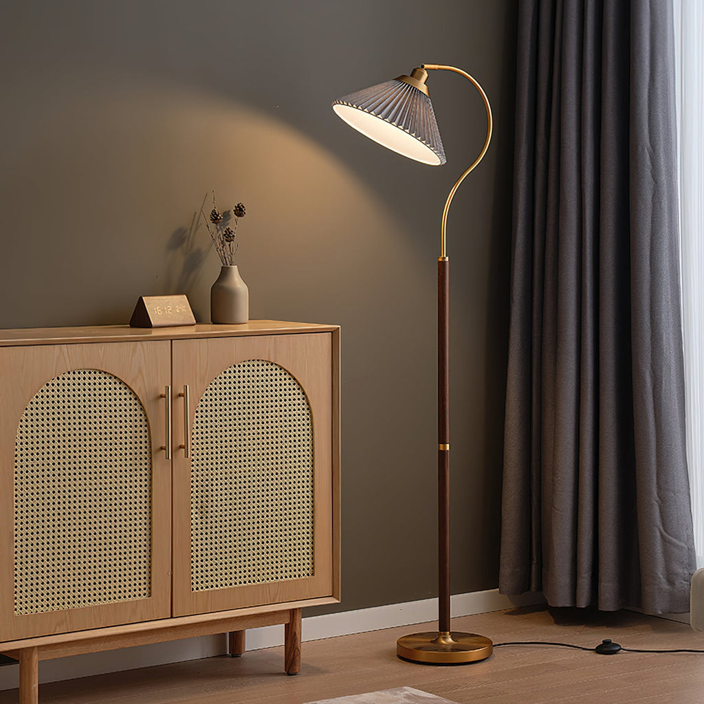 The grey pleated shade of this Clérachic floor lamp hangs on its curved brass arm beside a cane-fronted oak cabinet and a dark curtain.