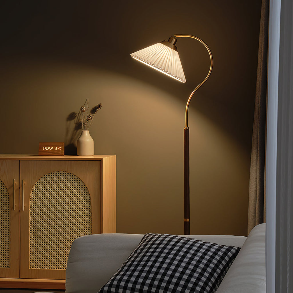 Seen from a low angle, a Clérachic floor lamp with a white pleated shade lights a taupe wall above a cane cabinet and a checked cushion.
