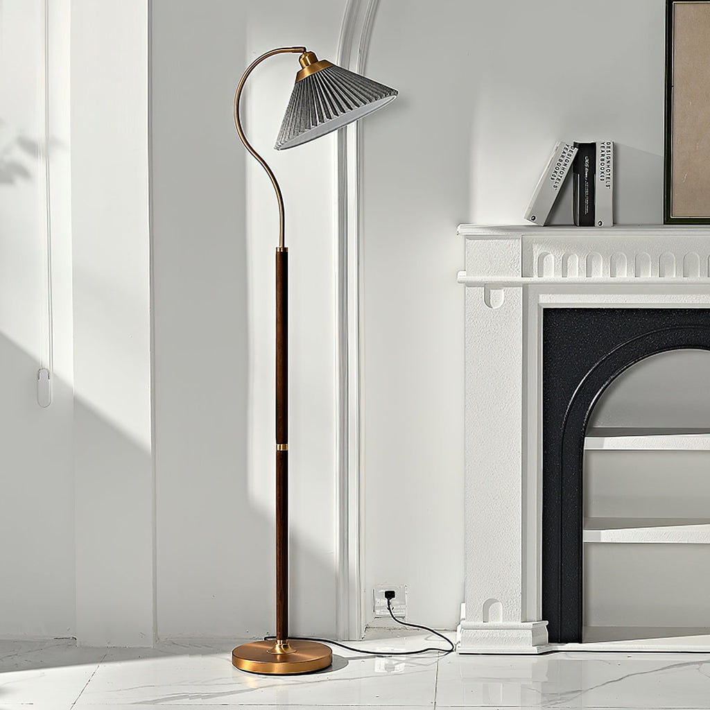A Clérachic floor lamp with a grey pleated shade stands by a white fireplace with books on the mantel, its cord running to a wall socket.