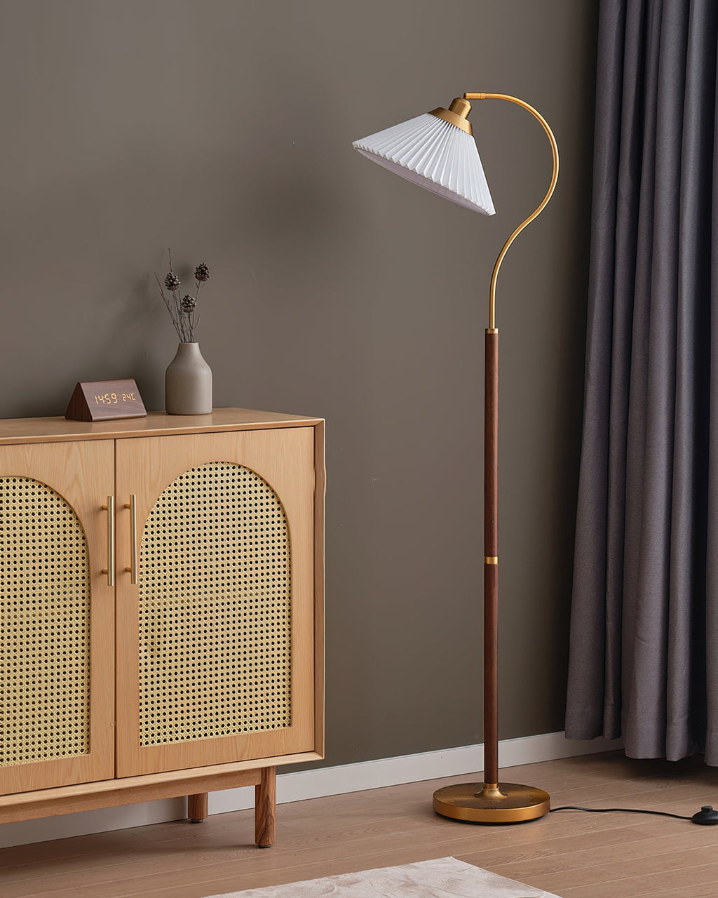 White pleated Clérachic floor lamp on a curved brass arm beside a cane-doored oak cabinet and heavy grey curtains, with its foot switch on the floor.