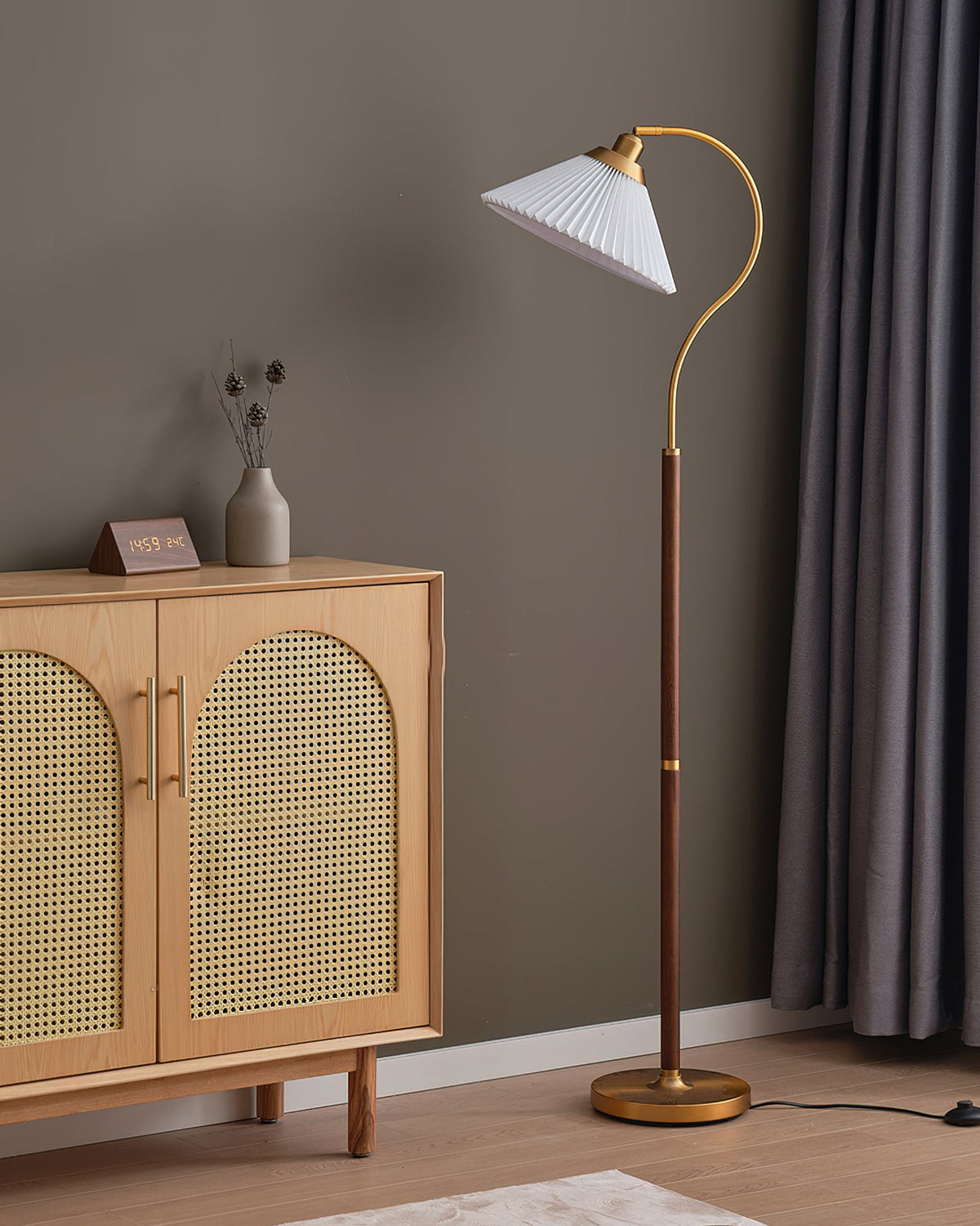 White pleated Clérachic floor lamp on a curved brass arm beside a cane-doored oak cabinet and heavy grey curtains, with its foot switch on the floor.
