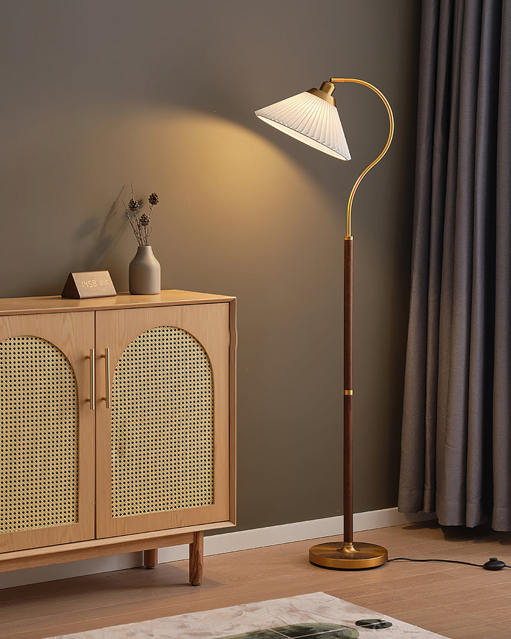 The Clérachic floor lamp with a white pleated shade, lit, throws a warm cone of light on the taupe wall above a cane cabinet.