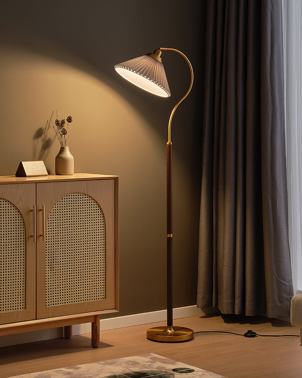 A lit Clérachic floor lamp with a grey pleated shade glows against a taupe wall, with a cane cabinet and a pleated curtain beside it.